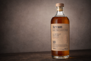 Bouteille d’Arran 10 ans photographiée seule sur une surface sombre, avec un fond taupe doux et une lumière latérale discrète.