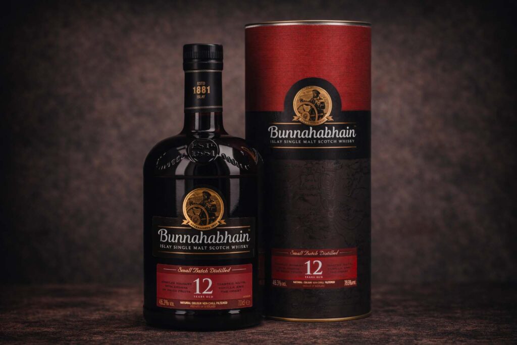 Bouteille de Bunnahabhain 12 ans accompagnée de son étui cylindrique rouge et noir, photographiés sur une surface sombre avec un fond texturé discret.