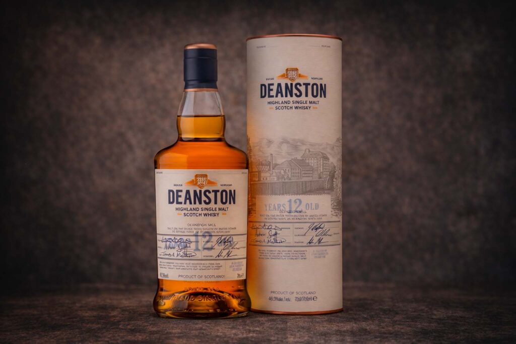 Bouteille de Deanston 12 ans accompagnée de son étui cylindrique crème, photographiés sur une surface sombre avec un fond texturé discret.